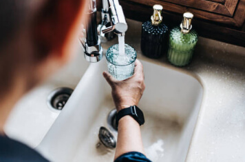 A person standing over a kitchen sink filling a clear glass with water.