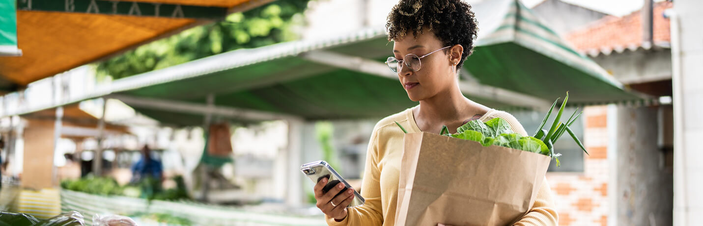 A woman with a paper bag full of groceries holding a cell phone